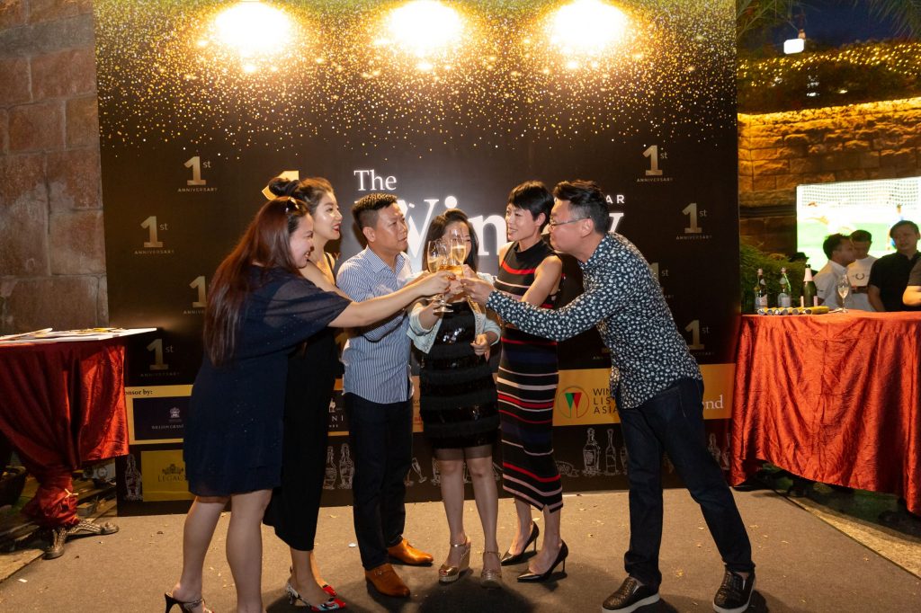 A group of six people are celebrating an occasion by toasting with champagne. They are outdoors at night, standing in front of a banner that reads "1st Anniversary". The banner has a sparkling gold and black design, indicating a festive event. Two of them are popping a champagne bottle with a spray of champagne visible in the air, while the others are holding their glasses up for a toast. They are all smiling and appear to be in a cheerful mood. There is a red-covered table to the right and a backdrop that suggests the event is quite elegant.